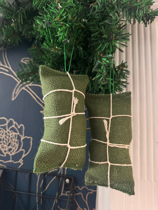 Two olive green burlap hanging ornaments shaped like wrapped pasteles, tied with white string, hanging from a green artificial pine tree branch.