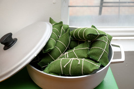 A pot with a lid, filled with green fabric-wrapped food parcels tied with white string, set on a green surface near a window.
