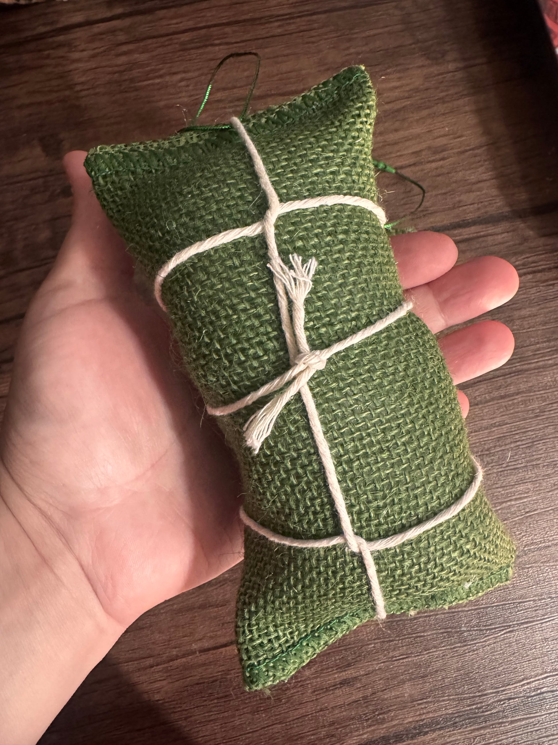 A small green burlap fabric pouch tied with white string, held in a person's hand over a dark wood surface.